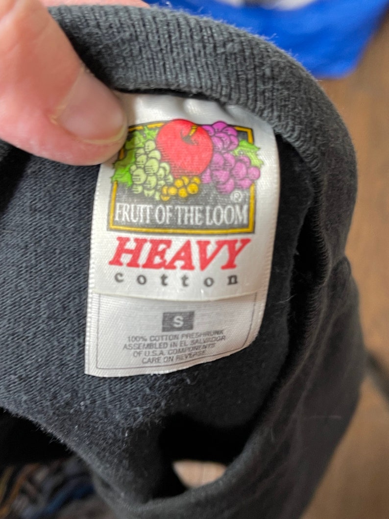 May include: Close-up of a black t-shirt tag. The tag features the "Fruit of the Loom" logo with a red apple, grapes, and other fruit. The tag reads "HEAVY cotton" and "S". The shirt is made of 100% cotton.