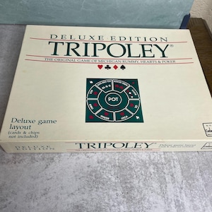 May include: A Deluxe Edition Tripoley game box. The box is cream-colored with red and black text. The game layout is printed on the box, with the words "Tripoley" and "Deluxe Edition" prominently displayed. The game is "The Original Game of Michigan Rummy, Hearts & Poker."