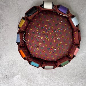 May include: A round, burgundy pin cushion with a floral pattern, surrounded by compartments holding spools of thread in various colors. Pins and needles are scattered across the cushion, with thread strands visible.