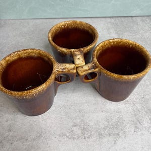 May include: Three brown ceramic mugs with a speckled glaze. Each mug has a rounded handle and a wide opening. The mugs are arranged in a triangular pattern, showcasing their design and color variations. The mugs are likely for coffee or tea.