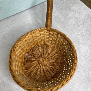 May include: A round, light brown woven wicker basket with a single long, cylindrical handle extending from one side. The basket features a circular base with a tight weave pattern that transitions into a wider, open lattice design toward the rim.