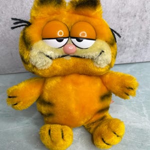 May include: A plush Garfield toy featuring bright orange fur with dark brown stripes, large plastic eyes with white and orange irises, a small pink nose, and black whiskers. The stuffed animal has a rounded body, short arms, and feet.