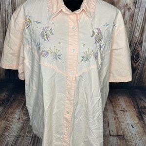 May include: A light peach-colored button-down shirt with short sleeves. The shirt features embroidered aquatic designs, including fish, seashells, and bubbles. The collar is embellished with small, white beads. The shirt is displayed on a black mannequin.