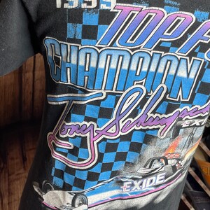 May include: Black t-shirt featuring a racing design with the text "1999 Top Fuel Champion Tony Schumacher Exide". The graphic includes a checkered flag pattern and a race car image. The shirt is made of a soft material.