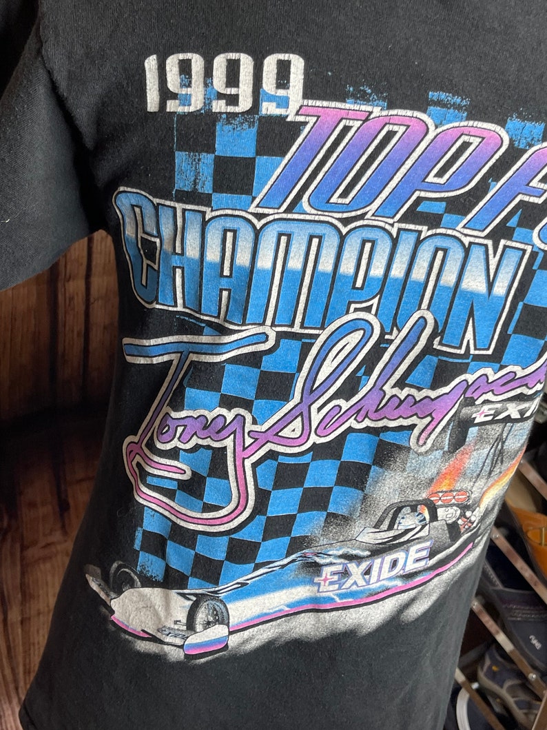 May include: Black t-shirt featuring a racing design with the text "1999 Top Fuel Champion Tony Schumacher Exide". The graphic includes a checkered flag pattern and a race car image. The shirt is made of a soft material.
