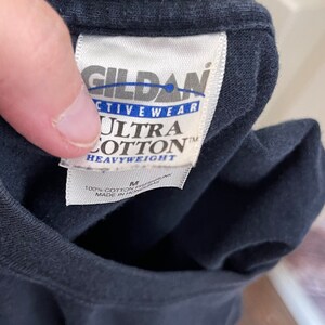 Może przedstawiać: Czarny t-shirt z białą metką Gildan Activewear Ultra Cotton Heavyweight. Metka zawiera nazwę marki, rozmiar M, informację o składzie 100% bawełny oraz kraj produkcji Honduras. Tkanina jest w jednolitym czarnym kolorze.