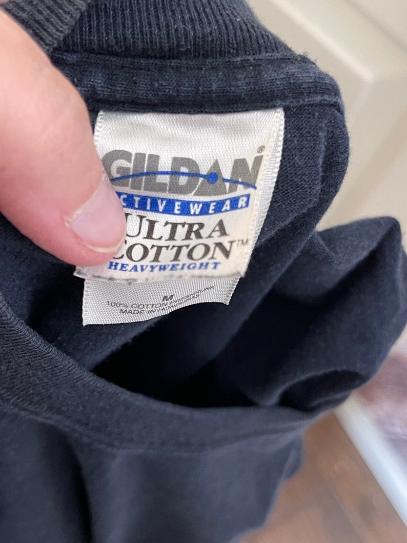 Może przedstawiać: Czarny t-shirt z białą metką Gildan Activewear Ultra Cotton Heavyweight. Metka zawiera nazwę marki, rozmiar M, informację o składzie 100% bawełny oraz kraj produkcji Honduras. Tkanina jest w jednolitym czarnym kolorze.