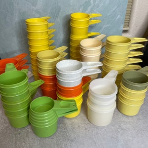 May include: A collection of vintage measuring cups in various colors, including yellow, green, orange, white, and brown. The cups are stacked and arranged in a group on a light blue surface.