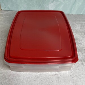 May include: A rectangular food storage container with a clear base and a red lid. The lid is flat with a slightly recessed center. The container is made of plastic and is designed for storing food.