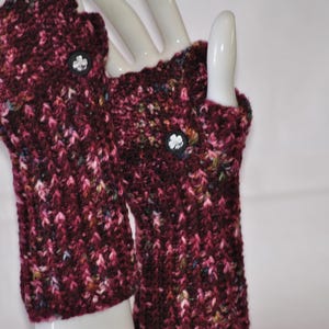 May include: A pair of burgundy fingerless gloves with a textured knit pattern. Each glove features a small, black button with a white floral design. The gloves are displayed on a white mannequin hand.