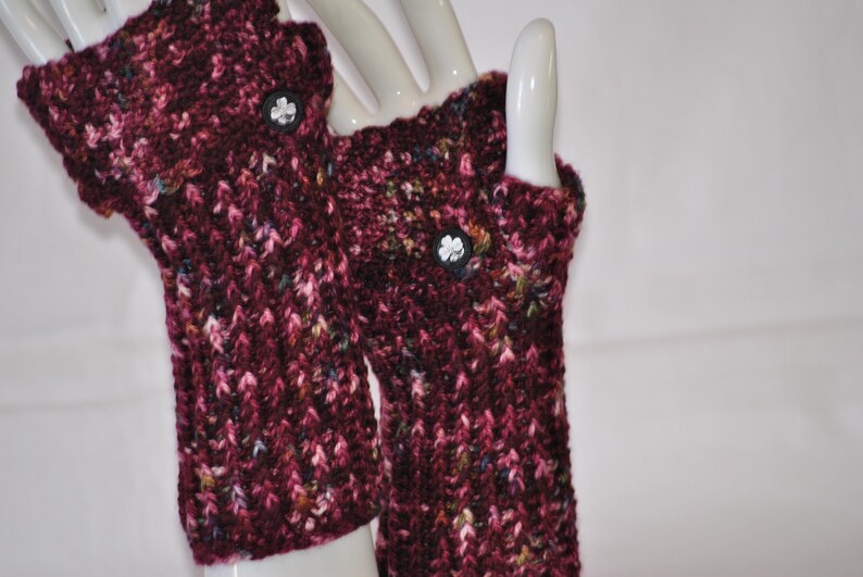 May include: A pair of burgundy fingerless gloves with a textured knit pattern. Each glove features a small, black button with a white floral design. The gloves are displayed on a white mannequin hand.