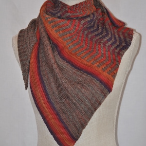 Puede incluir: Un chal triangular de punto con un degradado de color marr&oacute;n, naranja y morado. El chal se muestra en un maniqu&iacute; blanco.