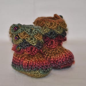 May include: A pair of handmade crochet baby booties featuring a variegated yarn in shades of red, orange, green, and brown. The cuffs are decorated with layered, scale-like crochet petals and small, iridescent dark beads along the top edge.