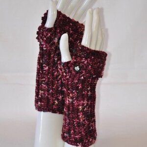 May include: A pair of burgundy fingerless gloves with a textured knit pattern. The gloves are displayed on white mannequin hands. A small, decorative button adorns the wrist of one glove. The gloves are a warm accessory.