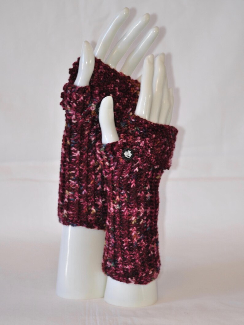 May include: A pair of burgundy fingerless gloves with a textured knit pattern. The gloves are displayed on white mannequin hands. A small, decorative button adorns the wrist of one glove. The gloves are a warm accessory.