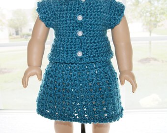 May include: A teal crochet doll outfit, consisting of a short-sleeved top with three pearl buttons and a matching skirt. The outfit is handmade with a textured stitch pattern, creating a unique and detailed look. The doll is positioned in front of a window.