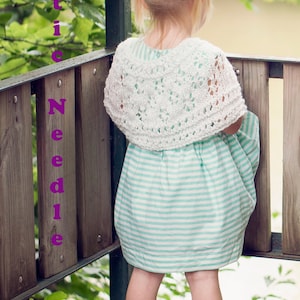 May include: A young girl wearing a white crocheted shawl over a light blue and white striped dress. She has her hair in pigtails and is standing on a wooden deck with a green railing.