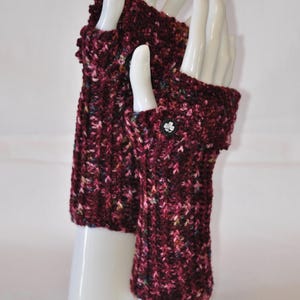 May include: A pair of burgundy fingerless gloves, displayed on white mannequin hands. The gloves are knitted with a textured pattern and feature a small, decorative button on the cuff. The yarn has flecks of pink, green, and yellow.
