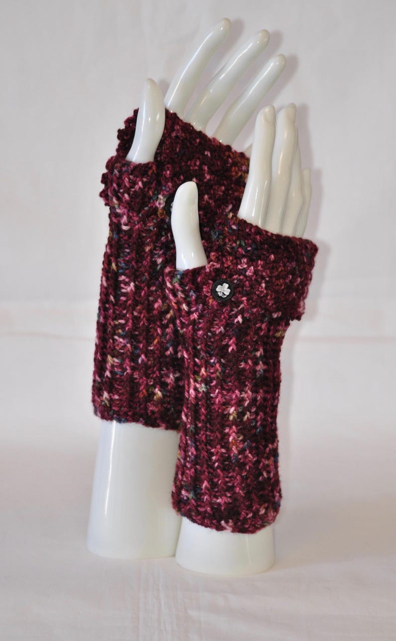 May include: A pair of burgundy fingerless gloves, displayed on white mannequin hands. The gloves are knitted with a textured pattern and feature a small, decorative button on the cuff. The yarn has flecks of pink, green, and yellow.