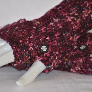 May include: A pair of burgundy fingerless gloves with a textured knit pattern. Each glove features a small, black button with a white floral design. The gloves are displayed on a white mannequin hand.
