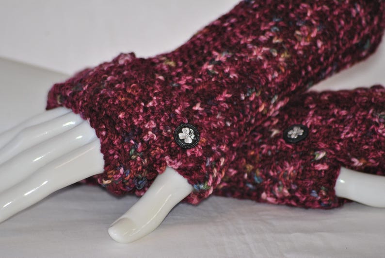May include: A pair of burgundy fingerless gloves with a textured knit pattern. Each glove features a small, black button with a white floral design. The gloves are displayed on a white mannequin hand.
