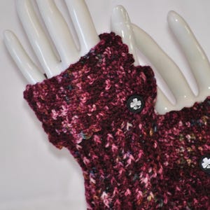 May include: Burgundy fingerless gloves with a textured knit pattern. The gloves feature a scalloped edge and are adorned with black buttons with a white clover design. The gloves are displayed on a white hand.