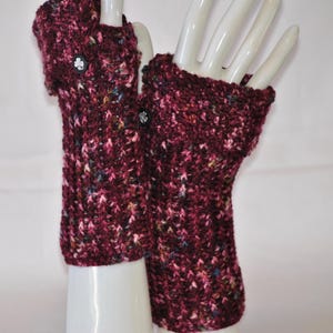 May include: A pair of burgundy fingerless gloves with a textured knit pattern. Each glove features a black button with a white floral design. The gloves are displayed on white mannequin hands.