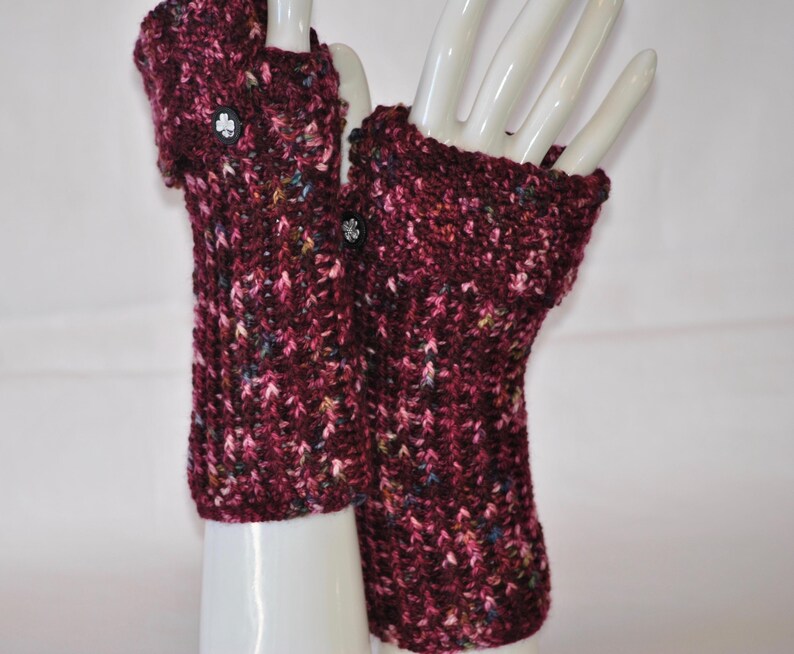 May include: A pair of burgundy fingerless gloves with a textured knit pattern. Each glove features a black button with a white floral design. The gloves are displayed on white mannequin hands.