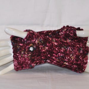 May include: A pair of burgundy fingerless gloves, hand-knitted with a textured yarn. The gloves feature a button closure and are displayed on a white hand form. The yarn includes flecks of various colors.