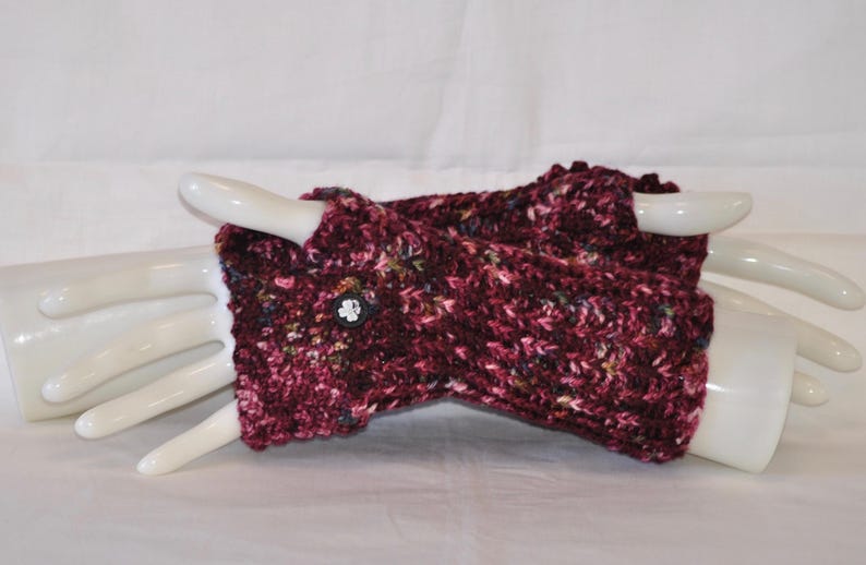 May include: A pair of burgundy fingerless gloves, hand-knitted with a textured yarn. The gloves feature a button closure and are displayed on a white hand form. The yarn includes flecks of various colors.