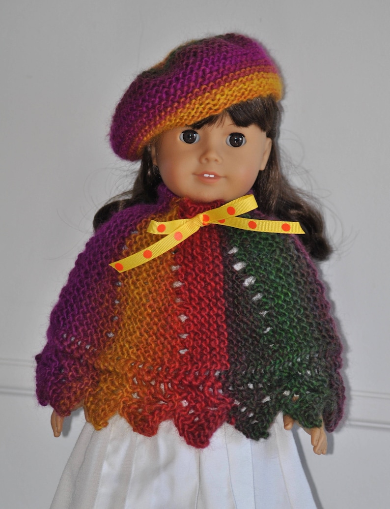 May include: A doll wearing a multi-colored knitted poncho with a yellow ribbon and polka dots, and a purple and orange knitted beret.