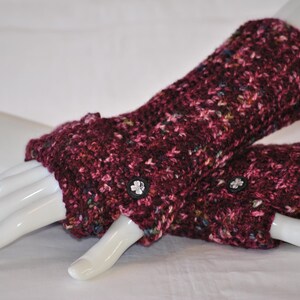 May include: A pair of burgundy fingerless gloves with a speckled pattern. The gloves are made of a thick, knitted material and feature small, decorative buttons. The gloves are displayed on a white mannequin hand.