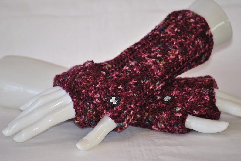 May include: A pair of burgundy fingerless gloves with a speckled pattern. The gloves are made of a thick, knitted material and feature small, decorative buttons. The gloves are displayed on a white mannequin hand.