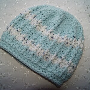 May include: A light blue crocheted hat with white stripes. The hat is made with a textured stitch and has a soft, cozy look.