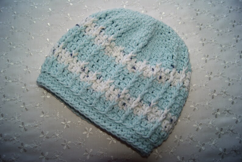 May include: A light blue crocheted hat with white stripes. The hat is made with a textured stitch and has a soft, cozy look.