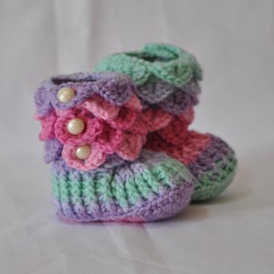May include: A pair of handmade crochet baby booties in pastel colors. The booties feature a layered design with pink, purple, and green hues, and are adorned with pearl-like buttons. The booties are designed to keep baby's feet warm.