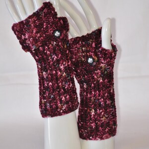 May include: A pair of burgundy fingerless gloves with a textured knit pattern. Each glove features a small, black button with a white clover design. The gloves are displayed on white mannequin hands.