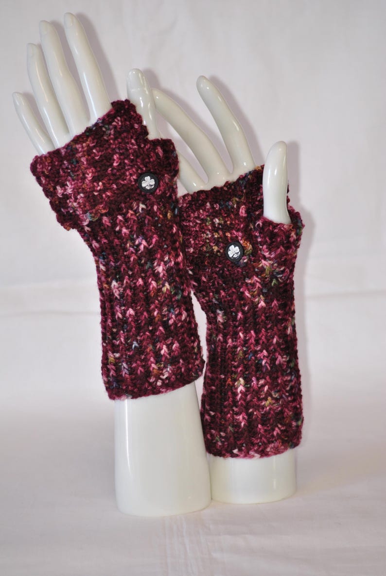 May include: A pair of burgundy fingerless gloves with a textured knit pattern. Each glove features a small, black button with a white clover design. The gloves are displayed on white mannequin hands.
