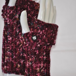 May include: A pair of burgundy fingerless gloves, hand-knitted with a speckled yarn. The gloves feature a button with a flower design on the cuff. The yarn includes flecks of pink, blue, and yellow. The gloves are displayed on a white mannequin hand.