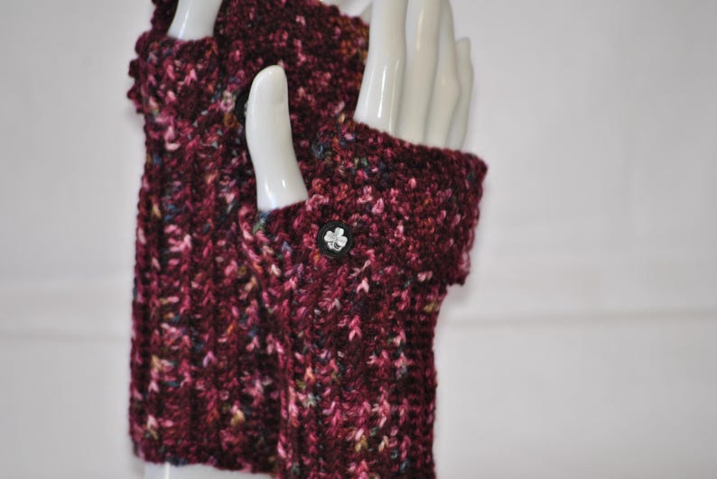 May include: A pair of burgundy fingerless gloves, hand-knitted with a speckled yarn. The gloves feature a button with a flower design on the cuff. The yarn includes flecks of pink, blue, and yellow. The gloves are displayed on a white mannequin hand.