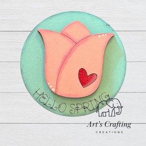 Puede incluir: Un letrero circular de madera con fondo verde menta claro que presenta un gran tulipán estilizado de color melocotón en el centro. Un pequeño corazón rojo está en el pétalo del tulipán. El texto HELLO SPRING está escrito en cursiva negra.