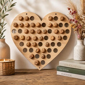 May include: A heart-shaped light wood display board featuring numerous circular cutouts designed to hold wine corks. The board is populated with various corks, each marked with unique black ink designs, symbols, and letters, arranged in a grid pattern.