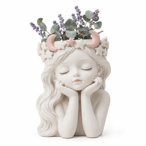 Może przedstawiać: Biała ceramiczna doniczka w kształcie postaci z zamkniętymi oczami, opierającej brodę na dłoniach. Głowa jest ozdobiona koroną z małych białych gwiazdek i dwóch różowych półksiężyców. W górnej części doniczki posadzono zielone rośliny.