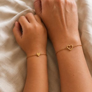 Könnte beinhalten: Zwei goldfarbene Kettenarmbänder an Handgelenken. Ein Armband hat einen kleinen, massiven Herzanhänger, während das andere Armband einen etwas größeren, offenen Herzanhänger besitzt. Beide Armbänder bestehen aus feinen, dünnen Metallgliedern.