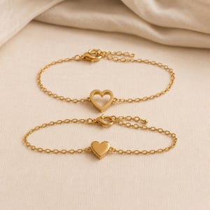 May include: Two gold-toned chain bracelets featuring heart-shaped charms. The top bracelet displays an open-frame heart charm, while the bottom bracelet features a solid, smooth heart charm. Both bracelets include a delicate link chain and a clasp.