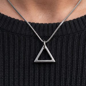 May include: A silver-toned necklace featuring a thin snake chain and a hollow triangular pendant. The metal has a matte, brushed finish. The pendant hangs from a simple jump ring attached to the chain, resting against a black textured jumper.