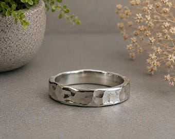 May include: A silver-toned ring featuring a hammered, textured surface with irregular facets. The band has a wide, substantial profile with a polished finish that reflects light across its uneven, metallic exterior surface.