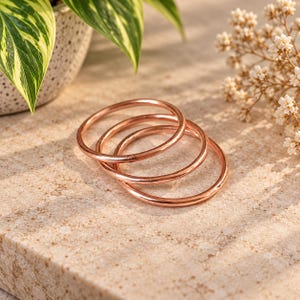May include: A set of three thin, circular copper-toned metal bangles with a smooth, polished finish. The bracelets are stacked slightly overlapping each other, showcasing a consistent metallic sheen and a simple, unadorned rounded band design.