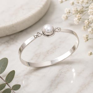 Op de afbeelding: Een zilverkleurige metalen armband met een enkele ronde witte parel in een ronde zetting. De parelzetting is aan beide zijden verbonden met de gladde, gepolijste band door middel van twee kleine ronde schakels, wat een doorlopend ontwerp vormt.