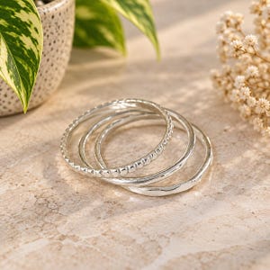 Puede incluir: Un conjunto de tres anillos de tono plateado apilados. Un anillo presenta una delicada textura de cuentas a lo largo de la banda exterior, mientras que los otros dos anillos tienen un acabado liso y pulido con una forma orgánica irregular.